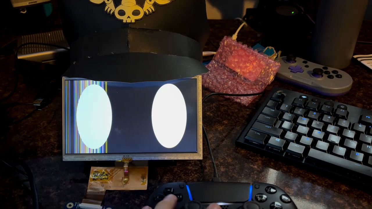 On a desk is a small LCD display with two ovals representing eyes drawn on it. On top of it sits a black papercraft captain hat with a yellow skull symbol on it. There is an orange ribbon cable connected from the bottom of the display, and a game controller is plugged in a lit near the bottom of the frame.