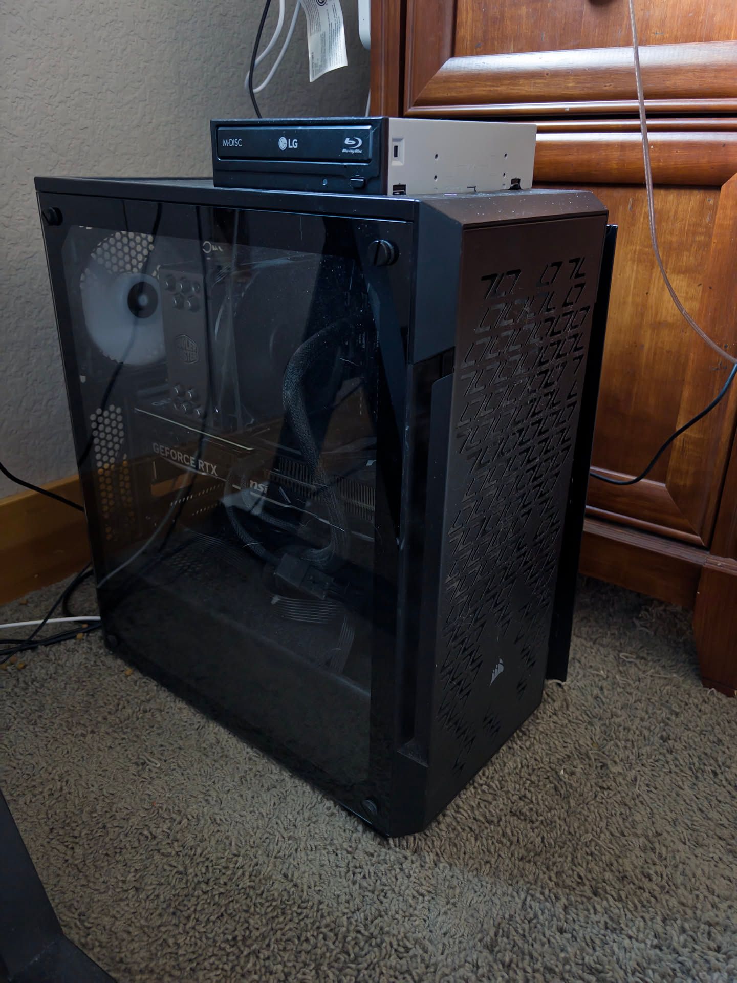 A computer tower. The case is made of black metal and plastic and has a glass side panel showing some of the internals, including a Cooler Master CPU fan and a GeForce RTX graphics card from MSI. On top of the tower, there is a Blu-ray Disc drive from LG labeled M-Disc. It faces to the side rather than the front. The tower sits on carpet with its back side facing a wall a few inches away. The side opposite the glass panel is a set of drawers. Some cables can be seen trailing behind the tower.